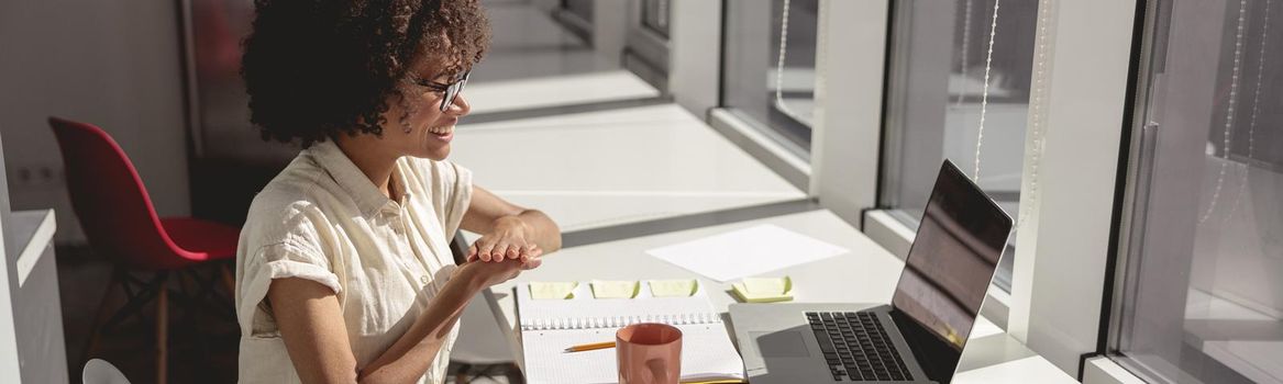 Smiling multiethnic woman learning and communicating in sign language online while sitting near window in the office