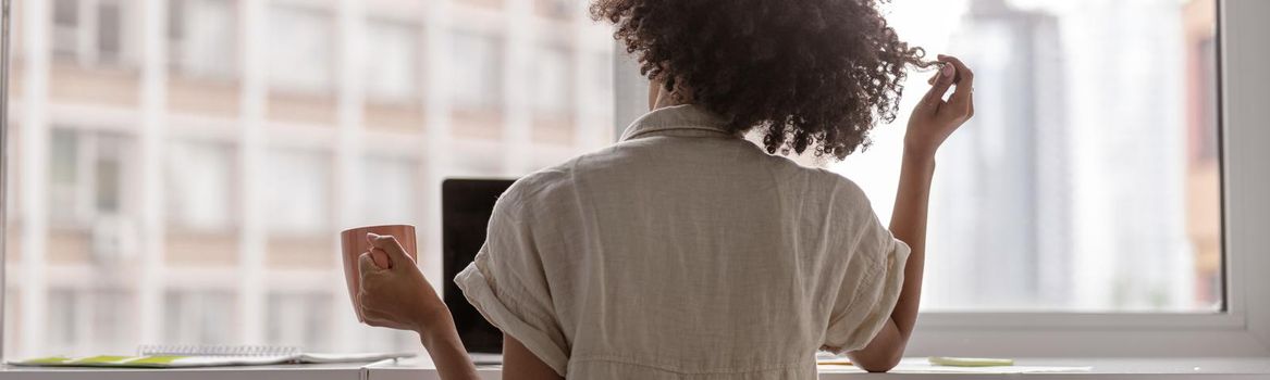 Back view of young woman sitting at desk near window while using laptop and holding cup. Copy space