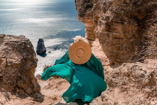 A beautiful young woman in a mint light dress with long legs stands on the edge of a cliff above the sea waving long dress, against the background of the blue sky and the sea