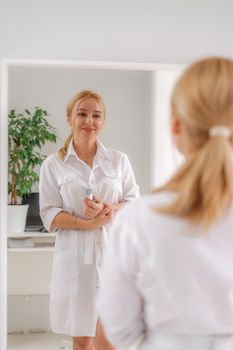 A blonde woman in white formal clothes looks at herself in the mirror. On a white background, green leaves at the back