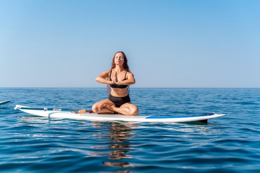 Sporty girl on a glanders surfboard in the sea on a sunny summer day. In a striped swimsuit, he sits in the lotus position with his eyes closed