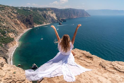 Portrait of a young woman on the beach by the sea, sitting with raised hands in a white dress and hat. Back photo.