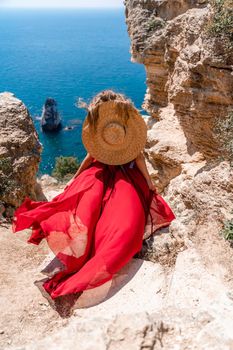 A girl with loose hair in a long red dress descends the stairs between the yellow rocks overlooking the sea. A rock can be seen in the sea. Sunny path on the sea from the rising sun.