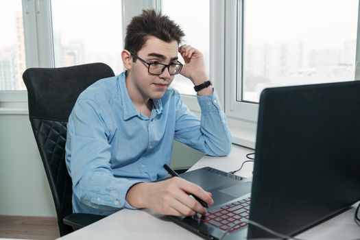 caucasian teenager guy thought about solving a problem while sitting at his workplace with a laptop and a graphic tablet