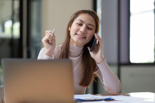 Portrait of young asian woman sitting at her desk working with calculator and paperwork while calling mobile phone in modern office