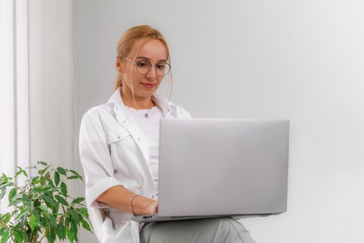 A blonde doctor in glasses or a cosmetologist in a white coat is sitting at a computer. She reads and types on the computer. Concept of medicine, beauty, cosmetologist, masseur