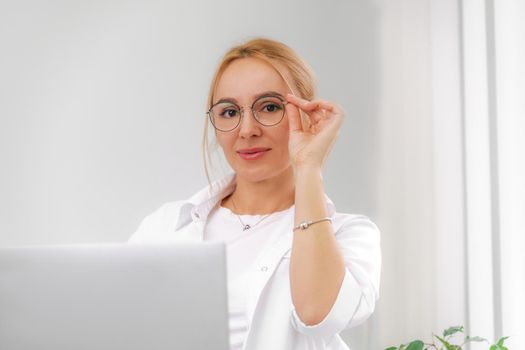 A blonde doctor in glasses or a cosmetologist in a white coat is sitting at a computer. She reads and types on the computer. Concept of medicine, beauty, cosmetologist, masseur