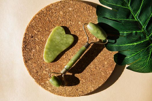 Jade Gua sha scraper and face roller massager on a cork round stand with a monstera leaf. Hard light, shadows, the concept self-care. Facial care. Zero waste. Lifting and toning treatment at home.