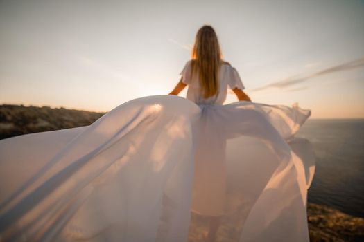 A beautiful young woman in a white light dress with long legs stands on the edge of a cliff above the sea waving a white long dress, against the background of the blue sky and the sea