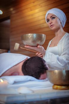 A woman performs Tibetan singing bowl therapy with a man lying under a white sheet. Relaxing meditation.