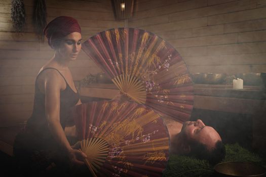A woman performs a healing ritual with a Chinese fan for a man. A man lies on spruce branches.