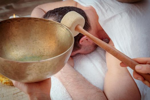 A woman performs Tibetan singing bowl therapy with a man lying under a white sheet. Relaxing meditation.