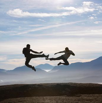 Two male kickboxers fly kicking one another on a mountain top.