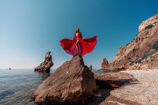 Beautiful woman posing in a luxurious red dress for the beach and in a white swimsuit with a long train, standing on a cliff against the sea. Around the cliff, the seashore