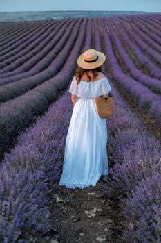 A middle-aged woman, blonde in a white dress and hat, walks through a lavender field with a basket.