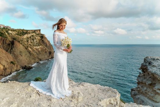 Summer bouquet of field daisies in the hands of a bride in a white dress against the backdrop of the sea. She stands on a rock above the sea. Copy space. The concept of calmness, silence and unity with nature