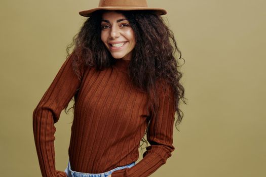 Enjoyed stylish curly Latin female in brown hat, smiling at camera, holding hand in jeans, saying Yeah, isolated green background. Copy space clothing fashion brands, free place for your ad.