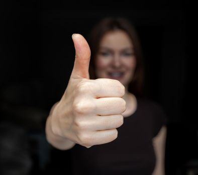 A closeup image of a young woman doing and showing a thumbs-up on a dark background. The concept of success
