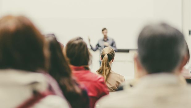 Male speaker giving presentation in lecture hall at university workshop. Audience in conference hall. Rear view of unrecognized participant in audience. Scientific conference event.