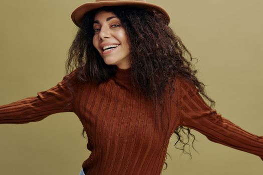Enjoyed stylish curly Latin female in brown hat, smiling at camera, gesturing Come on, saying Yeah, isolated green background. Copy space clothing fashion brands, free place for your ad.
