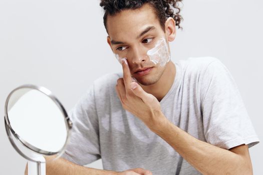 guy with curly hair applying cream on the face and looking in the mirror cosmetology light background. High quality photo