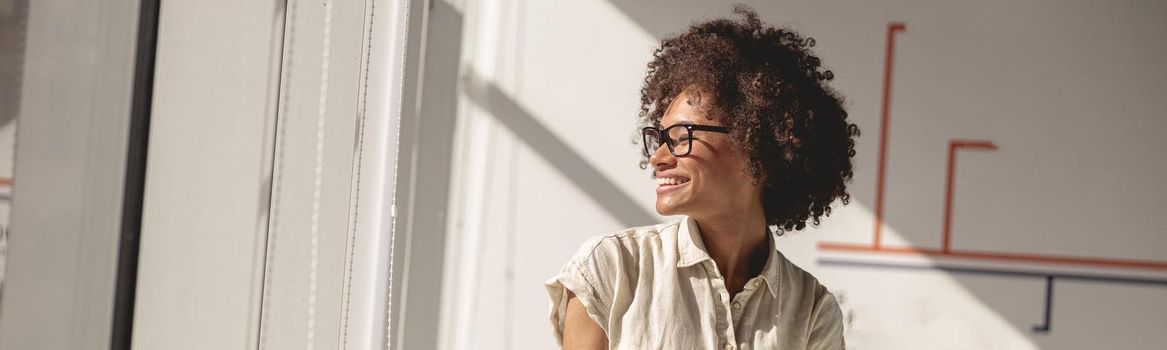 Happy Afro American female worker sitting on the table while holding notebook and looking out the window