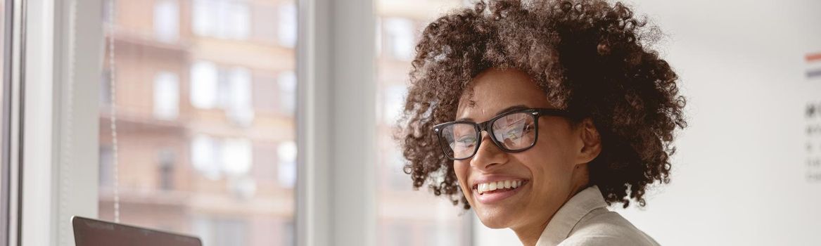 Smiling pretty African American lady wearing glasses and working with paper and laptop
