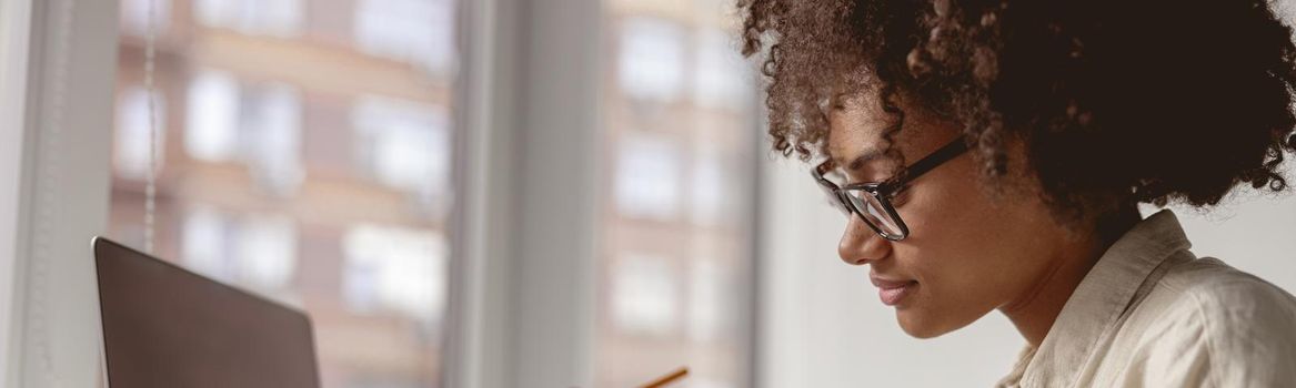 Pretty Afro American woman working with laptop and looking on document while sitting near window