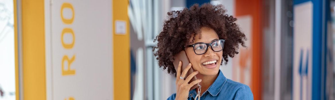 Portrait of smiling Afro American woman in glasses standing in office corridor while talking on smartphone