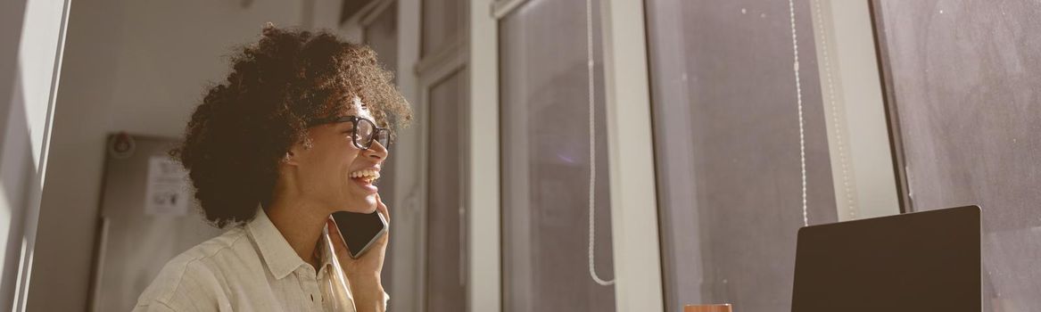 Smiling young female worker sitting at workplace in coworking while using smartphone