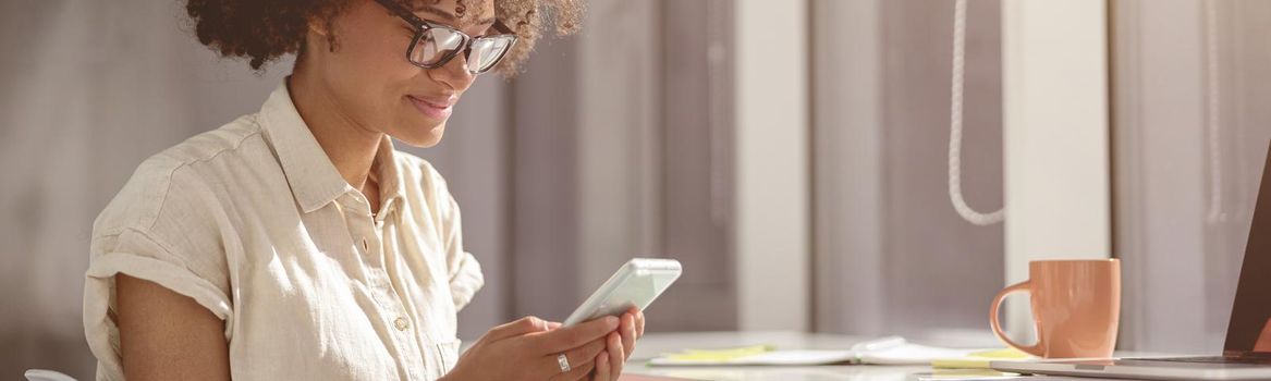 Pretty multiethnic lady wearing glasses and holding smartphone and looking at screen in the office