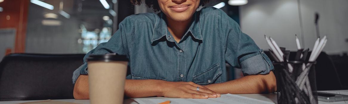 Smiling attentive woman wearing glasses and sitting at workplace with cup of coffee and document at the table