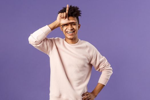 Portrait of charismatic happy, boastful young hispanic guy mocking friend for losing to him, showing loser gesture over forehead and laughing over defeated person, stand purple background.