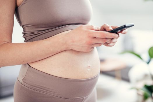 Closeup photo of pregnant female belly. Woman holding and using mobile smart phone app at home interiors. Pregnancy, technology, online shopping, preparation and expectation concept.