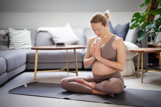 Young beautiful pregnant woman practising yoga at home in her living room. Motherhood, pregnancy, healthy lifestyle, well being and yoga concept