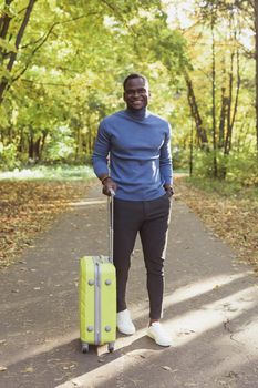 Cheerful african american young man in stylish clothes with suitcase walks in an autumn park on warm sunny day. Countryside travel concept