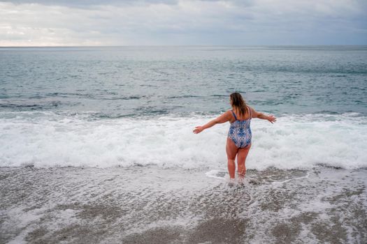 A plump woman in a bathing suit enters the water during the surf. Alone on the beach, Gray sky in the clouds, swimming in winter