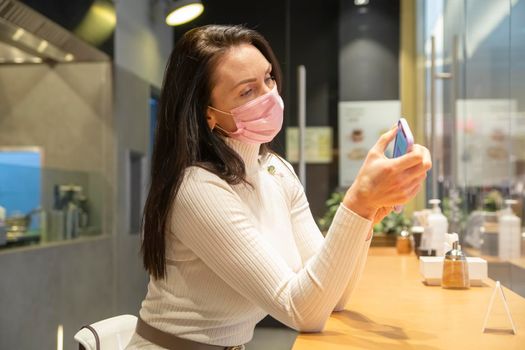 middle aged woman in medical mask in public place. portrait of woman in cafe typing smartphone