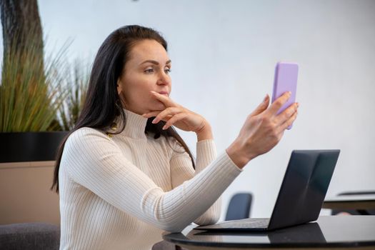 portrait of beautiful woman video calling via smartphone sitting by laptop