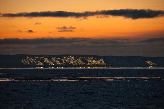 Long telephoto View of Collingwood and Blue Mountain and Craigleith Ski Hills Across Georgian Bay between sunset and twilight in winter. The ski hills are lit up with night lights and the sky is a rich sunset orange color and ice pack is gathered in the bay. High quality photo