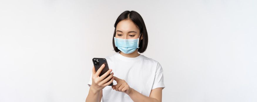 Health and covid-19 concept. Young asian woman in medical mask using mobile phone, typing on smartphone, standing against white background.