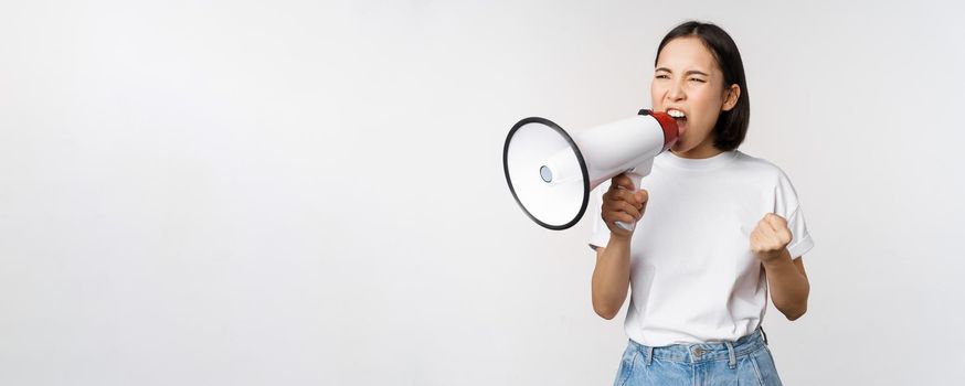 Confident asian woman shouting in megaphone, screaming and protesting. Girl activist using speaker to speak louder, standing over white background.