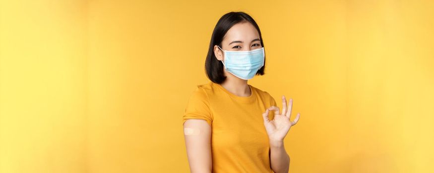 Vaccination from covid and health concept. Happy asian girl showing okay, wearing medical mask, band aid on shoulder, got coronavirus vaccine shot, yellow background.