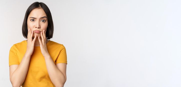 Shocked korean girl touching her face, looking concerned at camera, standing in yellow t-shirt over white background.