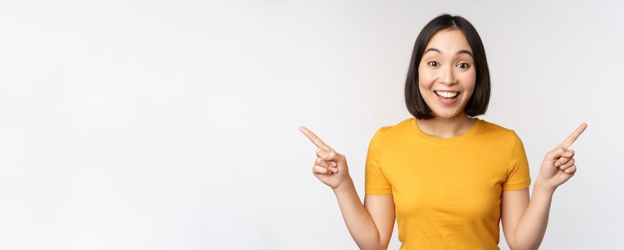 Cute asian girl pointing fingers sideways, showing left and right promo, two choices, variants of products, standing in yellow tshirt over white background.