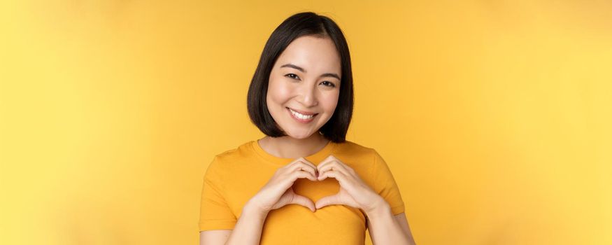 Close up portrait of smiling korean woman, showing romantic heart sign and looking happy, standing over yellow background. Copy space