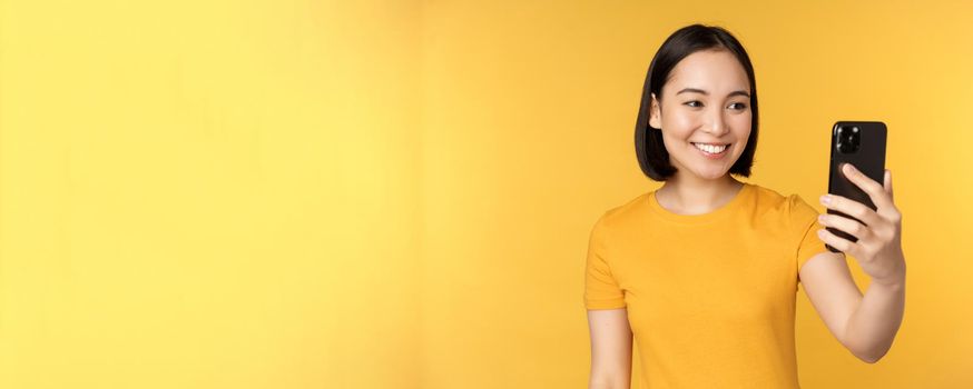 Image of happy, beautiful asian girl video chatting, talking on smartphone application, standing against yellow background. Copy space