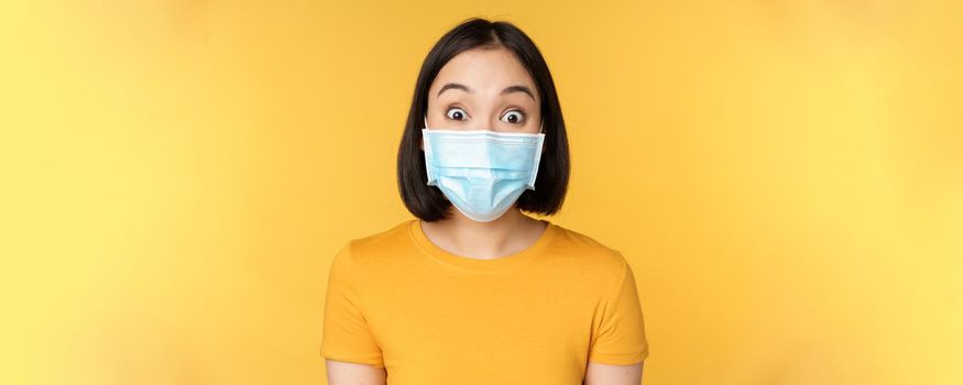 Covid-19 and medical concept. Close up portrait of asian woman in face mask, looking surprised and amazed at news, standing over yellow background.