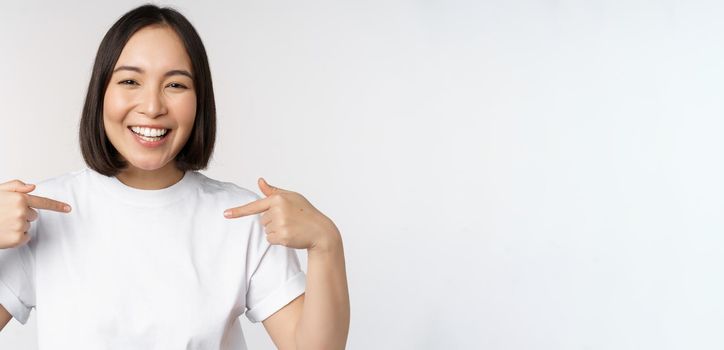 Happy and confident asian woman, student smiling and pointing at herself, self-promoting, showing logo on t-shirt, standing over white background.