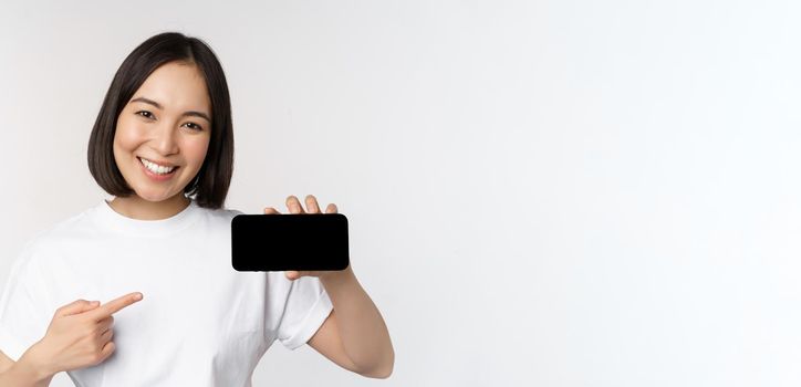 Portrait of smiling korean woman pointing finger at mobile phone screen, showing horizontal smartphone display, recommending website or store online, white background.